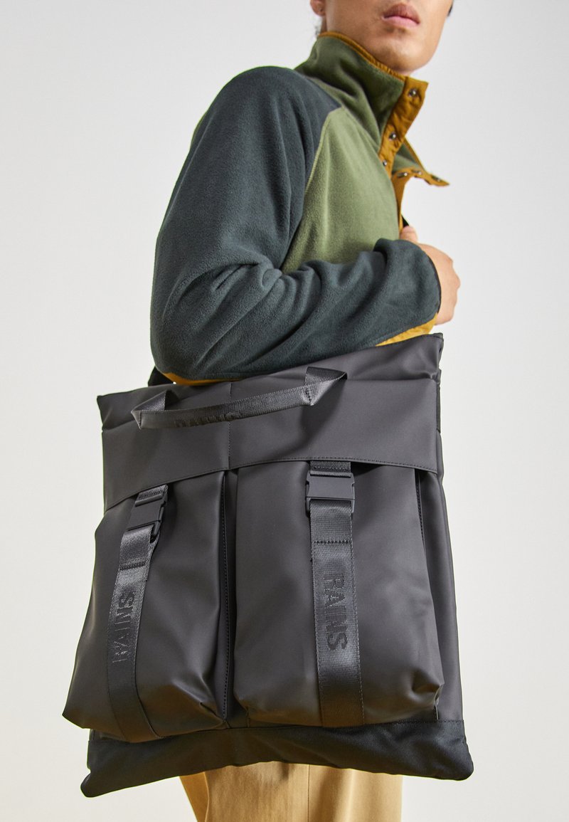 Black, waterproof tote bag with dual front straps, fabric handles, and a structured shape. Smooth texture with subtle branding.