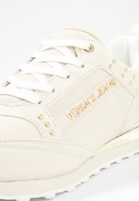 Baskets en daim blanc avec des accents dorés, présentant un panneau latéral estampillé, des lacets plats blancs et une semelle texturée.