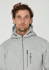 Sudadera gris con cremallera y capucha ajustable, con un bolsillo en el pecho con cremallera y un logo bordado en el lado izquierdo. Textura suave.