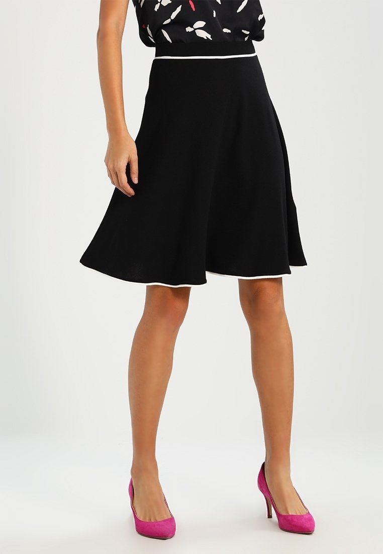 Femme portant une jupe noire mi-longue avec un bord blanc, un haut noir à motifs, et des chaussures à talons hauts roses vifs, debout sur un fond uni.
