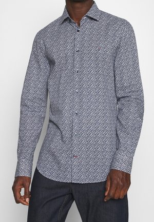 Man draagt een nauwsluitend overhemd met lange mouwen en knopen, met een klein wit bladmotief op een donkere achtergrond, gecombineerd met donkere jeans.