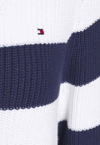 Pull tricoté blanc et marine à rayures, présentant une texture côtelée et une petite étiquette logo avec des accents rouges, blancs et bleus.