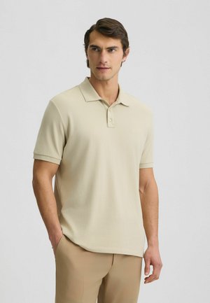 Polohemd in hellem Beige, kurze Ärmel, weicher Stoff, betont durch ein dezentes Logo auf der linken Brust, ergänzt durch hellfarbige Hosen.