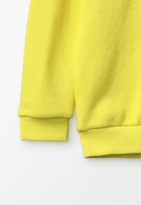 Žlutá mikina se strukturovaným pleteným vzorem, žebrovanými manžetami a volným střihem. Látka vypadá měkce a prodyšně.