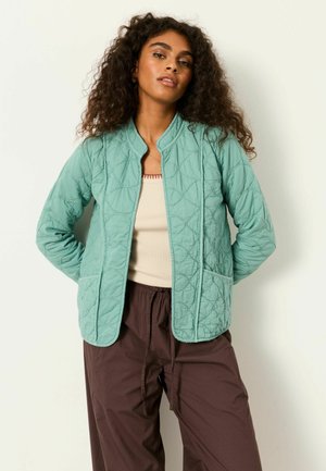 Frau mit lockigem Haar, die eine hellgrüne gesteppte Jacke, ein beiges Top und lockere braune Hose trägt, steht mit den Händen hinter dem Rücken.