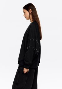 Sudadera negra de gran tamaño con tachuelas plateadas en las mangas, con cuello redondo y puños acanalados, combinada con vaqueros anchos de pierna oscura.