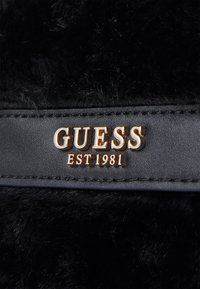 Materiale in pelliccia sintetica nera con una fascia in pelle liscia; logo dorato "GUESS" stampato in rilievo con "EST 1981" sottostante; enfasi sul contrasto di texture.