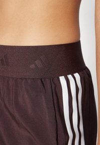 Shorts de sport marron avec une large taille élastique présentant un logo subtil. Des bandes blanches sur les côtés apportent un contraste au tissu texturé.