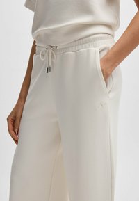 White sweatpants made of soft material with an elastic waistband, drawstring, side pockets, and subtle embroidered logo detail.