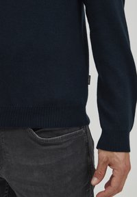 Sötétkék kötött pulóver bordázott szegéllyel és mandzsettával, oldalán egy kis címkével. Fekete farmernadrág felett viselve, kézi részletekkel.