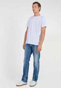 Camiseta de algodón blanca combinada con jeans de mezclilla azul claros. Modelo vistiendo zapatillas blancas con suela beige, posando de pie contra un fondo liso.