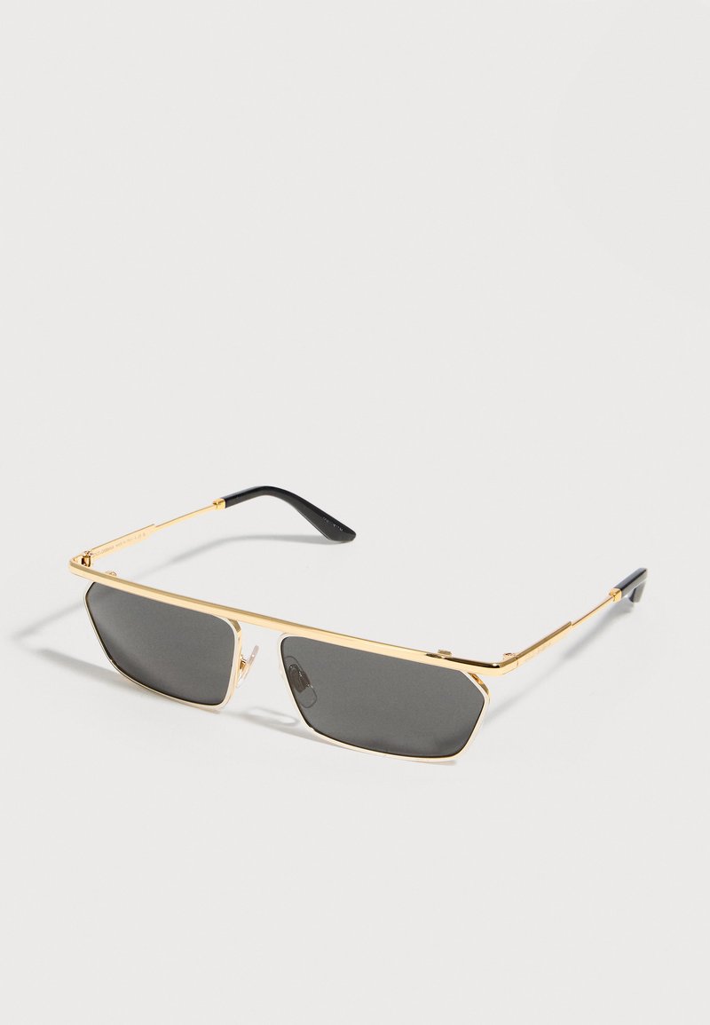 Schlanke rechteckige Sonnenbrille mit schwarzen Gläsern und dünnen goldfarbenen Metallrahmen, die auf einer weißen Oberfläche liegt.