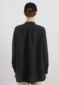 Zwarte langekorte mouw shirt gemaakt van lichtgewicht stof met een rechte snit, kraag en knoopmanchetten; gedragen met een zwarte honkbalpet.