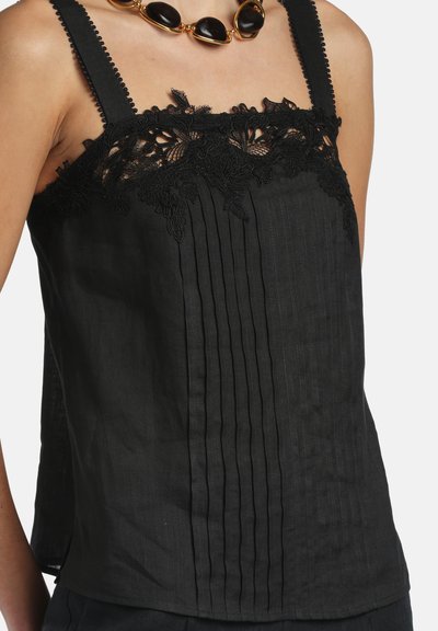 Femme portant un haut noir sans manches avec un bord en dentelle et des plis verticaux, associée à un collier épais en perles dorées et noires.