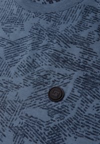 Modré tričko z texturované tkaniny s jemným tmavým vzorem a kulatým černým logem poblíž výstřihu.