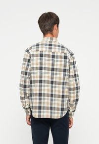Geruite shirt in beige, marineblauw en bruin. De stof is gestructureerd, met knoopsluitingen en een gebogen zoom. De achterkant is duidelijk zichtbaar.
