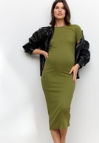 Olijfgroene geribbelde midi-jurk, nauwsluitend ontwerp, korte mouwen, aangevuld met een losse zwarte leren jas. Gladde textuur, zonder patronen.