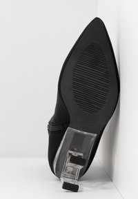 Soulier noir à bout pointu avec une semelle texturée et un talon bloc transparent, reposant contre une surface d'angle blanche.