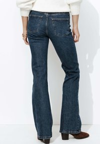 Jean évasé bleu foncé en denim, doté d'un design à cinq poches et de coutures visibles, associé à des bottes à talons marron.