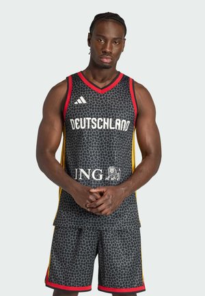 Mann in schwarzem und grauem Basketballtrikot und Shorts mit der Aufschrift "Deutschland" und Sponsorlogos, stehend mit verschränkten Händen.