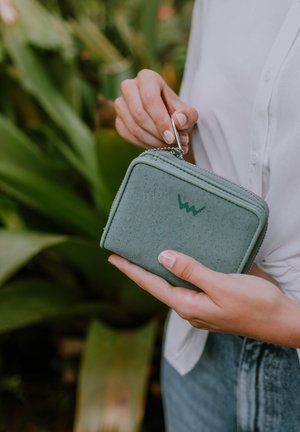 Green cork wallet, zippered closure, rectangular shape, textured surface, logo detail on the front, held by a hand against a foliage background.