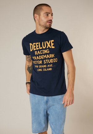 Homme portant un t-shirt graphique bleu marine "DEELUXE" et un short en denim bleu clair, regardant vers sa gauche sur un fond uni.