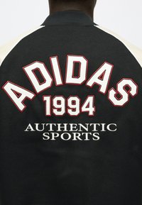 Bakside av en svart jakke med hvit og rødkantet tekst "ADIDAS 1994" og brodert "AUTHENTIC SPORTS" nedenfor.