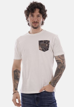 Uomo con capelli ricci che indossa una t-shirt bianca con taschino sul petto scuro a motivo e jeans, che mostra tatuaggi su entrambi gli avambracci.