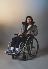 A person in a wheelchair wearing a brown puffer jacket, white t-shirt, dark pants, and blue sneakers against a soft gradient background.