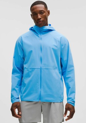 Młody mężczyzna ubrany w jasnoniebieską kurtkę z kapturem i jasnoszare szorty, stojący na jednolitym białym tle.