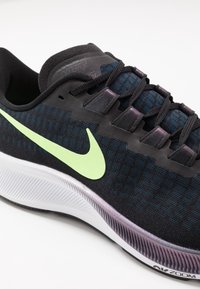 Musta Nike urheilujalkine, jossa on teksturoitu verkkomateriaali, vihreä swoosh-logo, purppurat yksityiskohdat ja valkoinen pehmustettu välipohja.