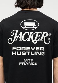 Άτομο που φοράει μαύρο μπλουζάκι με λευκό κείμενο "Jacker Forever Hustling MTP France" και γραφικό ενός πάγκου πάνω από το κείμενο.
