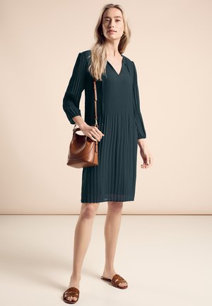 Dunkelgrünes Faltenkleid mit V-Ausschnitt, langen Ärmeln und A-Linie; kombiniert mit einer braunen Leder-Umhängetasche und braunen Sandalen.