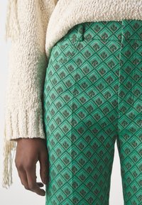 Gröna mönstrade byxor med en geometrisk diamantdesign och svarta accenter, kombinerat med en krämfärgad stickad tröja med flätade detaljer.