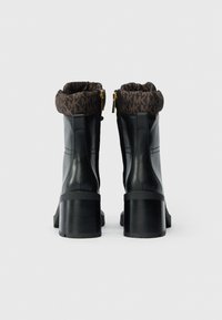 Botas de tornozelo em couro preto com um punho padrão castanho, fechos de correr dourados e saltos robustos, com textura suave e design estruturado.