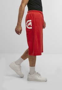 Red cotton shorts with a logo pocket, drawstring waist, knee-length, paired with white sneakers and light gray crew socks.