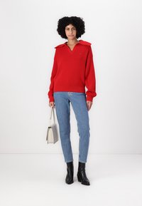 Rød ribbestrikket genser med krage, kombinert med lyseblå tettsittende jeans og svarte ankelstøvler. Modellen holder en lysegrå håndveske.