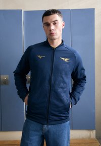 Veste zippée marine en tissu lisse avec un motif géométrique texturé ; accents de logo dorés. Portée avec un jean bleu clair.