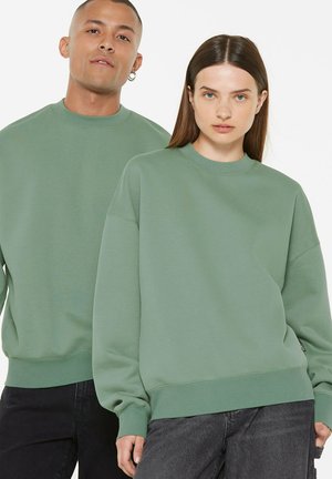 Two adults wearing matching green sweatshirts stand against a white background, facing forward with neutral expressions.