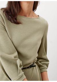 Oliwkowa bluzka z długim rękawem, z dekoltem w łódź i bufkami, zestawiona z czarnym paskiem z subtelnymi metalicznymi akcentami.