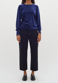 Marinblå sammet långärmad topp med samlad detalj vid axlarna och smockade ärmslut, kombinerad med svarta raka jeans och svarta loafers.