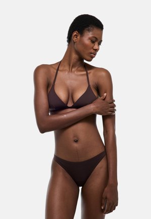 Femme aux cheveux courts portant un bikini marron foncé, debout avec un bras croisé sur le corps et regardant vers le bas sur le côté.