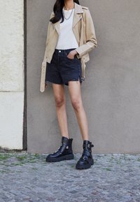 Chaqueta de moto de cuero beige sobre una camiseta blanca, combinada con pantalones cortos de mezclilla negros y botas de tobillo negras con hebillas y suelas gruesas.