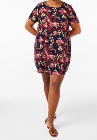 Kvinna iklädd en marinblå blommig klänning med rosa, beige och bruna mönster, tillsammans med bruna öppna sandaler med platt sula, som står med händerna bakom ryggen.