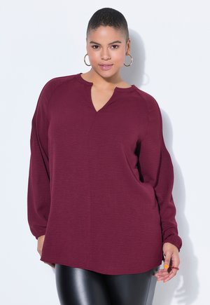 Lockige Frau mit goldenen Creolen, einer locker sitzenden weinroten Langarmbluse und schwarzen Leggings mit Lederoptik steht vor einer weißen Wand.