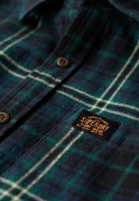 Flanellen overhemd in donker groen en marineblauw met een wit ruitpatroon; heeft bruine knopen en een zwart label met oranje tekst. Zachte textuur.