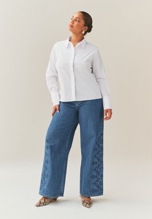 Femme en chemise blanche boutonnée et jean bleu à jambes larges avec un motif subtil sur les jambes, portant des chaussures pointues imprimé serpent, debout avec assurance.