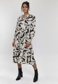 Abito lungo, bottonato, di color crema con motivo floreale nero, caratterizzato da un design a strati e maniche lunghe. Indossato con stivali neri alla caviglia.