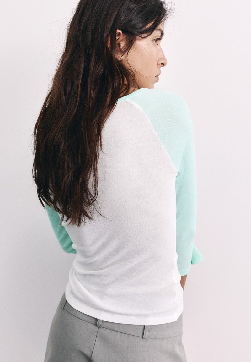 Femme aux longs cheveux foncés tournée de côté, portant une chemise blanche avec des manches bleu clair et un pantalon gris, sur un fond blanc uni.