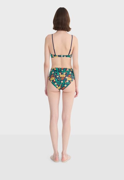 Femme debout, de dos, portant un bikini deux pièces avec un motif floral coloré et des fines bretelles noires sur le haut.